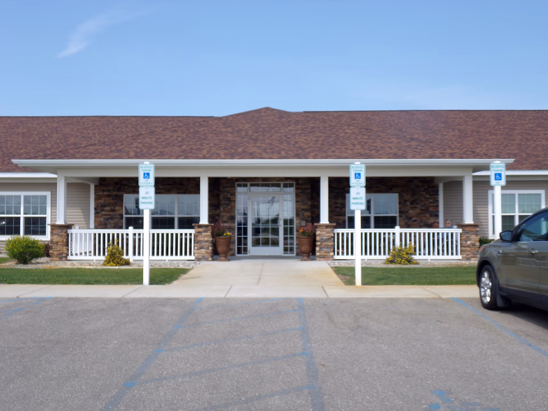 Front exterior view of Hale Creek Manor building with a covered entrance, stone and siding facade, white railings, and three reserved parking signs for handicapped parking in front.