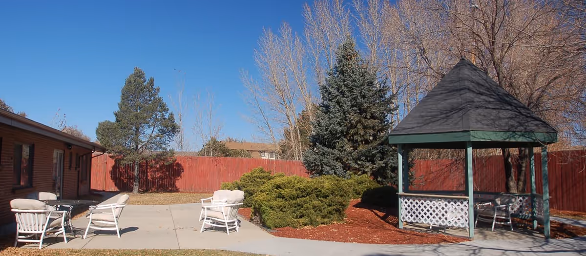 Outdoor courtyard with a gazebo, white chairs, red fence, and landscaped shrubs.