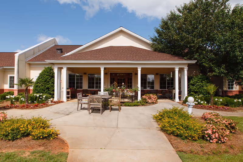 Front exterior view of The Brennity at Daphne Assisted Living & Memory Care building with a covered porch, outdoor seating area with wooden tables and chairs, surrounded by landscaped flower beds and greenery under a partly cloudy sky.