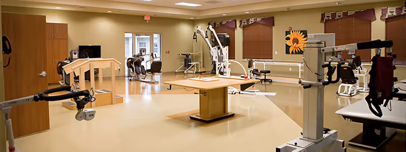 A spacious physical therapy or rehabilitation room with various exercise and therapy equipment including parallel bars, exercise bikes, and weight machines. The room has beige walls, a light tan floor, windows with brown blinds and valances, and a sunflower painting on the wall.