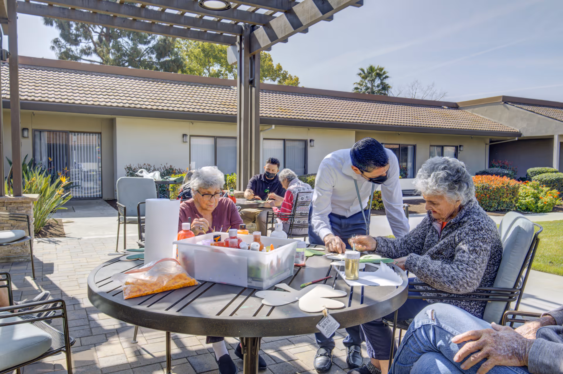 Several elderly individuals seated around a round outdoor table under a pergola, engaging in arts and crafts activities with paint bottles and paper. A staff member wearing a mask is assisting one of the elderly women. The background shows a single-story building with windows and a tiled roof, surrounded by greenery and a clear sky.