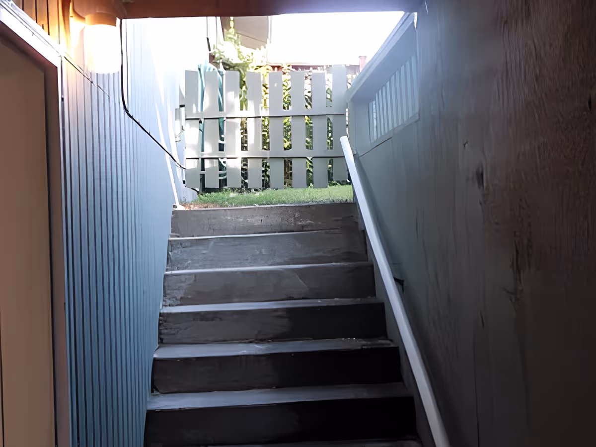 A concrete staircase leading upward to an outdoor area with a white wooden fence and green grass visible at the top. The stairwell walls are painted blue on the left and gray on the right, with a white handrail on the right side.