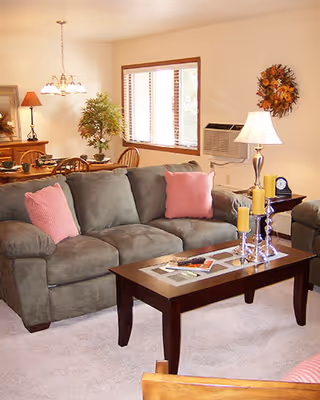 Bright living room with a green sofa, coffee table holding candles and magazines, a lamp, and a dining area visible in the background.