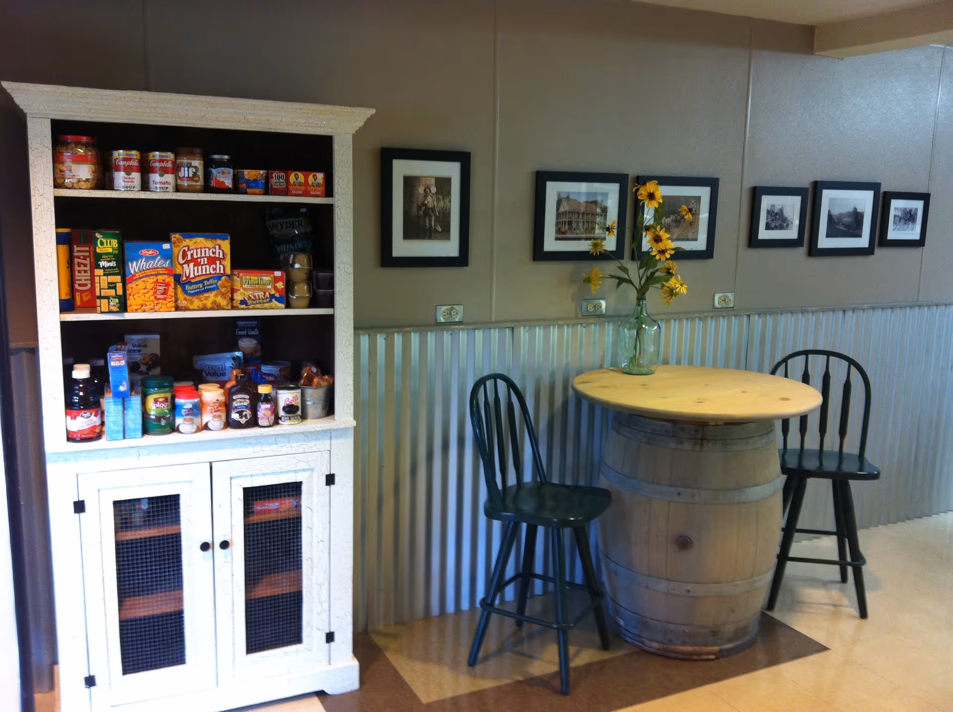 A cozy corner with a white wooden cabinet stocked with various snacks and food items on the left, and a round wooden table made from a barrel with two dark green chairs on either side. A vase with yellow flowers is placed on the table. The wall behind has framed black and white pictures and a corrugated metal wainscoting.