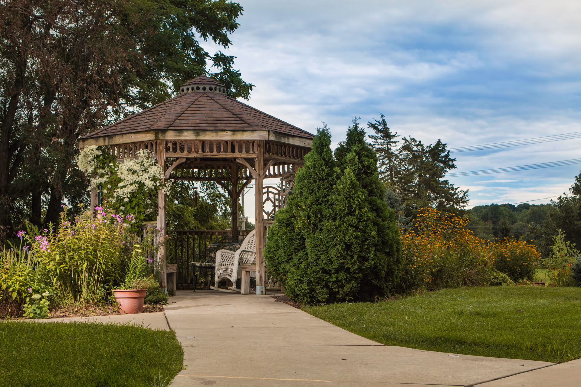 Wooden gazebo with white wicker chairs surrounded by flowering plants and greenery along a paved walkway.