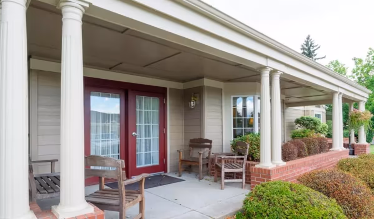 Covered porch area with white columns and brick base, featuring wooden chairs and a small table. Red double doors with glass panes and white curtains lead into the building. Shrubs and greenery surround the porch.