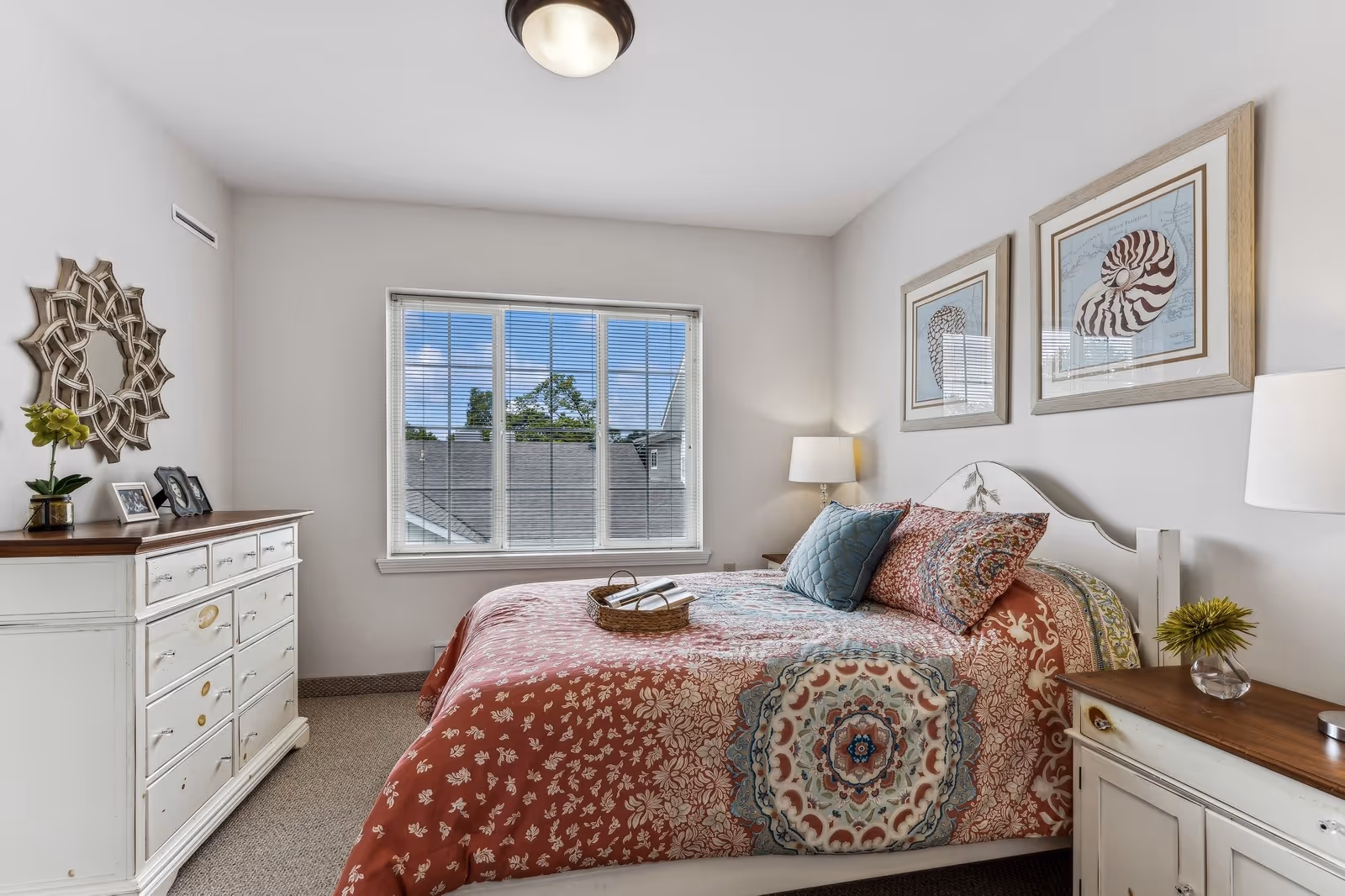A cozy bedroom with a large window letting in natural light. The bed has a colorful patterned quilt with multiple pillows. On either side of the bed are wooden nightstands with lamps and small decorative items. A white dresser with framed photos and a decorative mirror is against one wall. Two framed seashell prints hang above the bed.