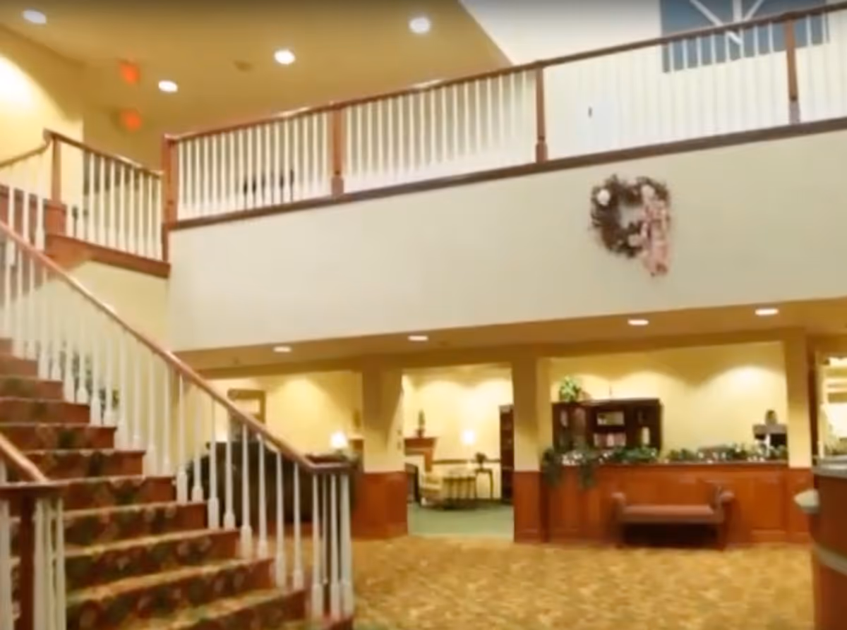 Interior view of a senior living facility lobby area with a carpeted staircase on the left, wooden railing on the upper level, a wreath decoration on the wall, and a seating area with plants and furniture under warm lighting.
