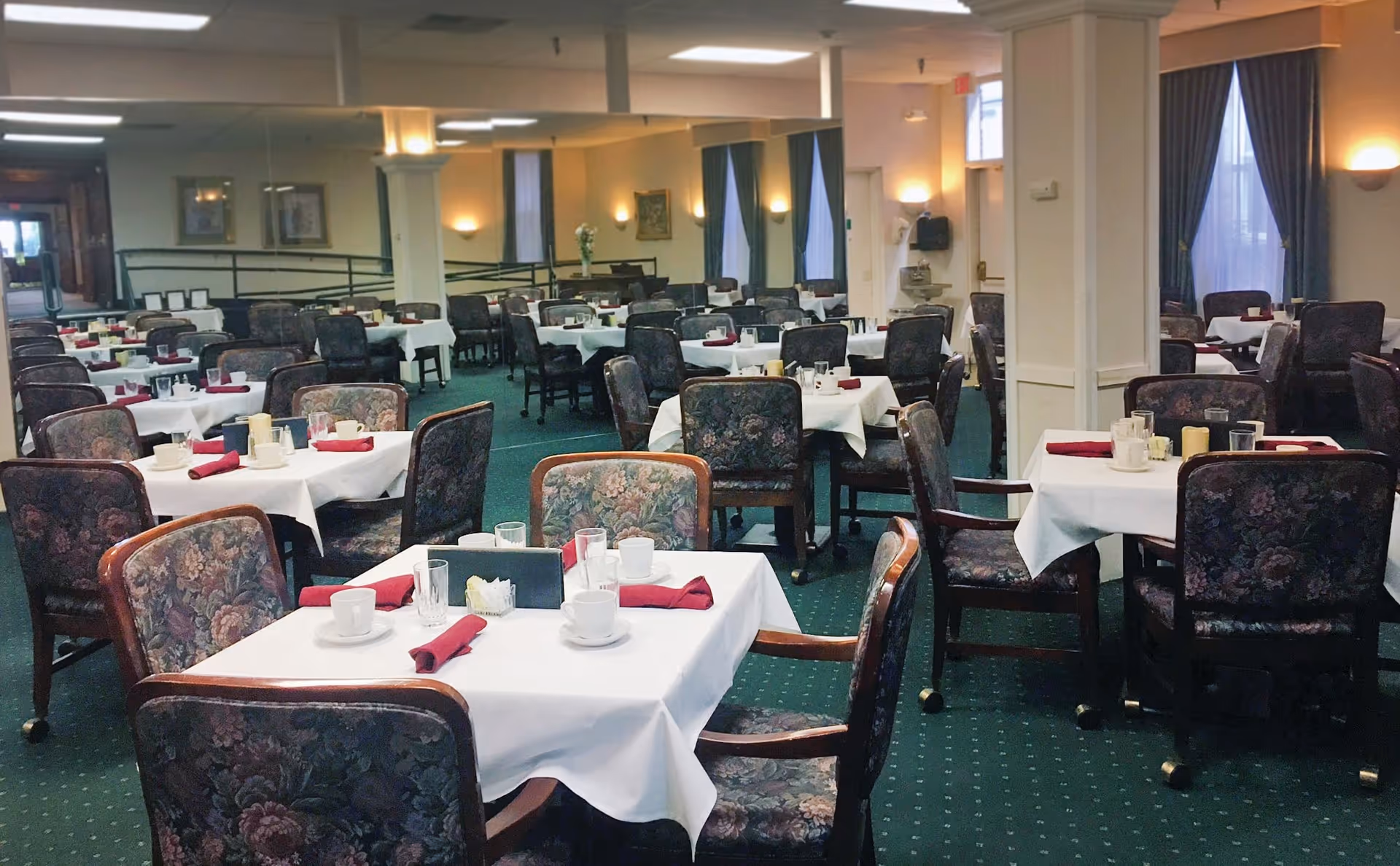 A spacious dining room with multiple tables covered in white tablecloths, each set with cups, glasses, napkins, and condiments. The chairs have floral upholstery, and the room is carpeted in green with soft lighting from wall sconces and ceiling lights. Large windows with blue curtains are visible along the walls.