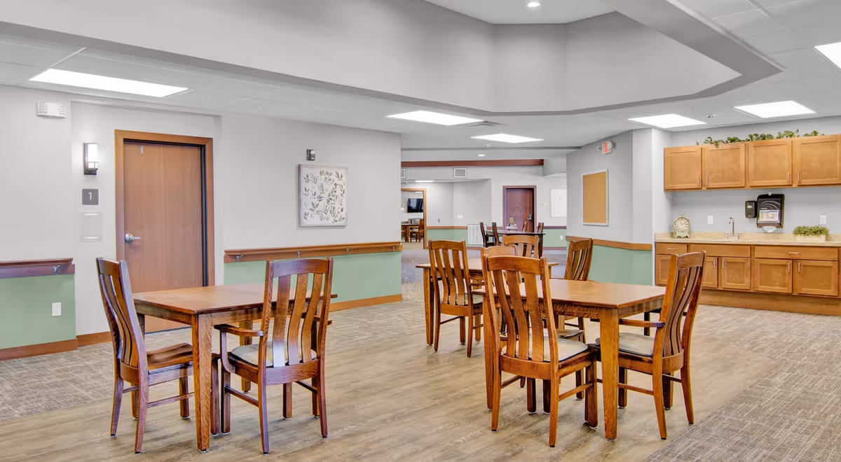 Open dining area in a senior living facility with wooden tables and chairs and a kitchenette along the back wall.