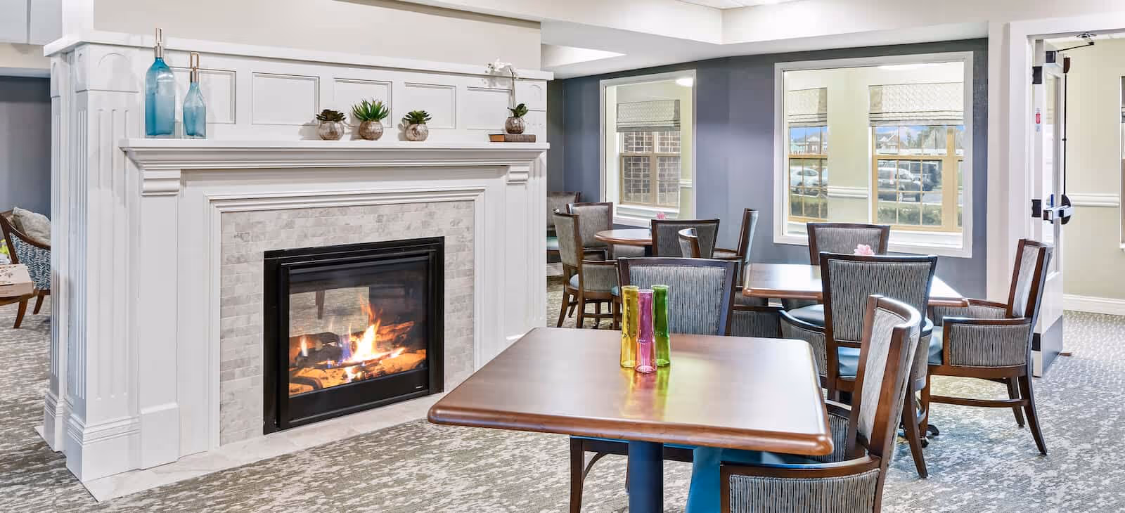 A cozy common area featuring a lit fireplace with decorative vases and small plants on the mantel. Several wooden tables and chairs are arranged around the room near windows that let in natural light.
