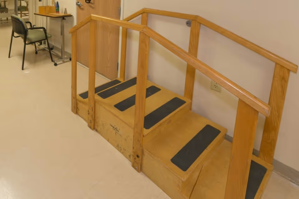 A small wooden rehabilitation step with handrails on both sides, placed indoors on a light-colored tiled floor. In the background, there is a chair and a table with some items on it, next to a closed wooden door.