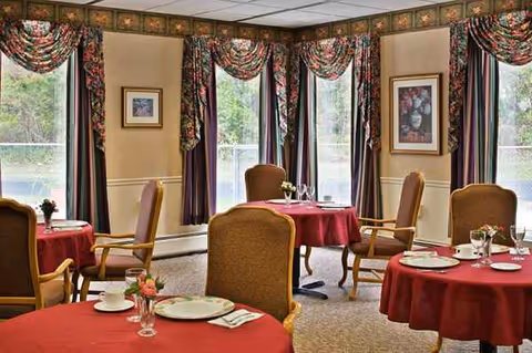 Senior living dining room with red tablecloths, upholstered chairs, and large windows.