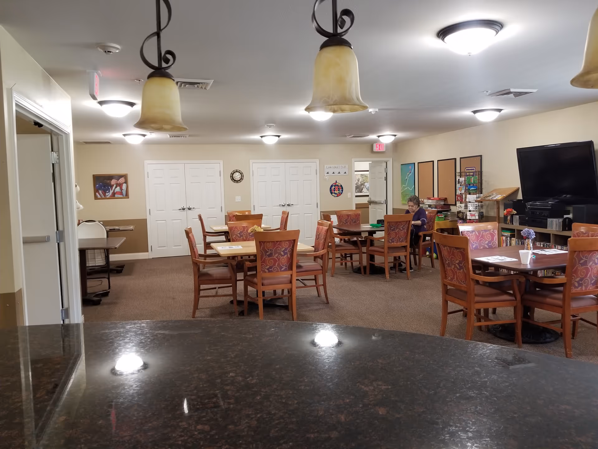 A common area in a senior living facility with several wooden tables and chairs arranged around the room. A single elderly woman is seated at one of the tables near the back, engaged in an activity. The room has beige walls, ceiling lights, hanging pendant lamps, a large TV on a stand, and various decorations and bulletin boards on the walls.