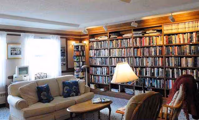 A cozy living room with a large bookshelf filled with books covering one wall. The room features a beige sofa with blue floral pillows, a wooden coffee table, a floor lamp, and several comfortable chairs. A window with sheer white curtains allows natural light to brighten the space.