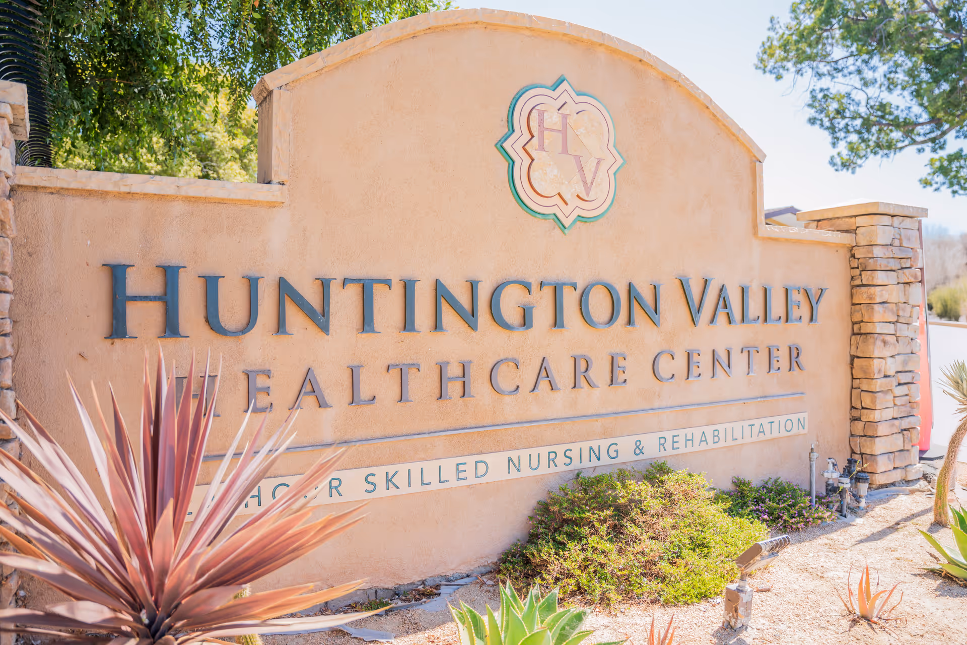 Stone sign for Huntington Valley Healthcare Center surrounded by desert landscaping with plants and trees in the background.