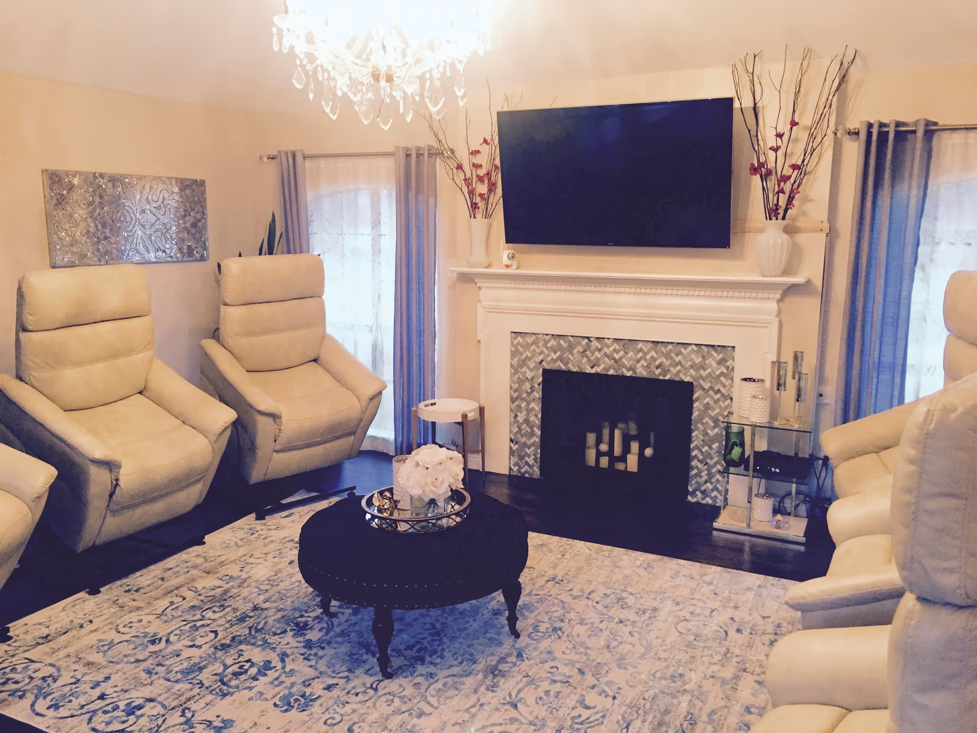 A cozy living room with four beige recliner chairs arranged around a round black ottoman with a floral centerpiece. The room features a fireplace with a decorative tile surround and a flat-screen TV mounted above it. Two windows with sheer curtains and blue drapes flank the fireplace. A chandelier hangs from the ceiling, and there is a patterned area rug on the floor.