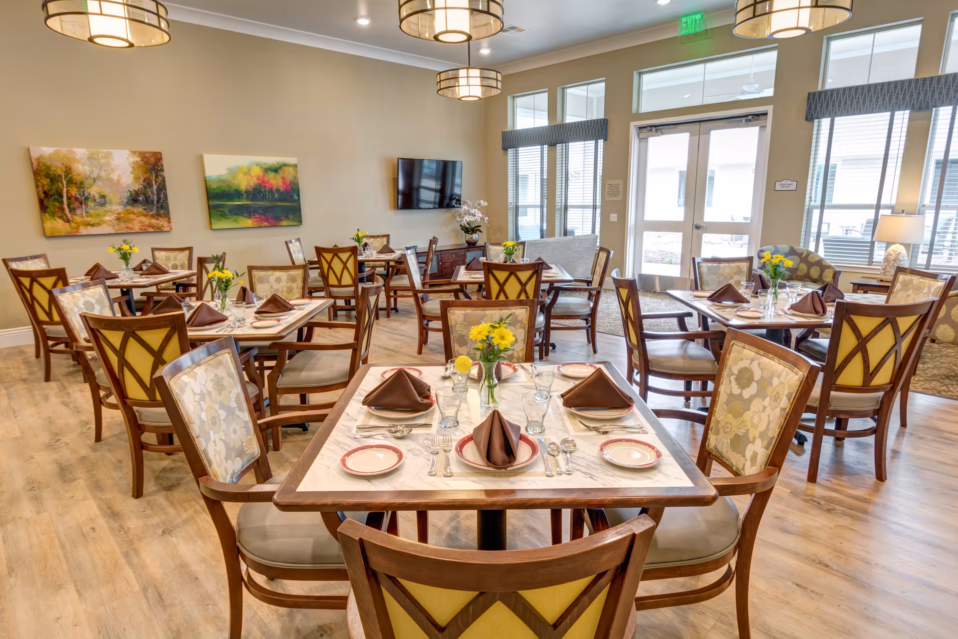 A bright and inviting dining room in a senior living facility with multiple tables set for four people each. The tables have white marble tops and are arranged with plates, glasses, silverware, and brown folded napkins. Yellow flowers in small vases decorate each table. The room features wooden chairs with patterned upholstery, large windows letting in natural light, two landscape paintings on the wall, a mounted flat-screen TV, and modern ceiling lights.