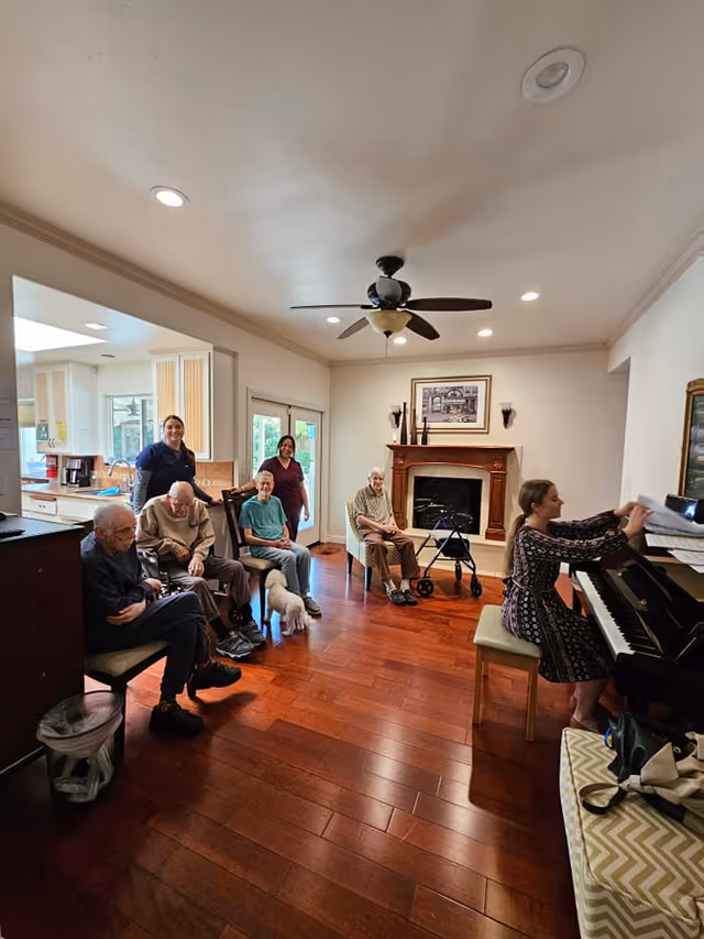 A group of elderly people sitting in a living room with two caregivers standing behind them. One elderly woman is sitting near a fireplace with a walker beside her. A woman is playing the piano on the right side of the room. The room has wooden floors, a ceiling fan, and a kitchen visible in the background.