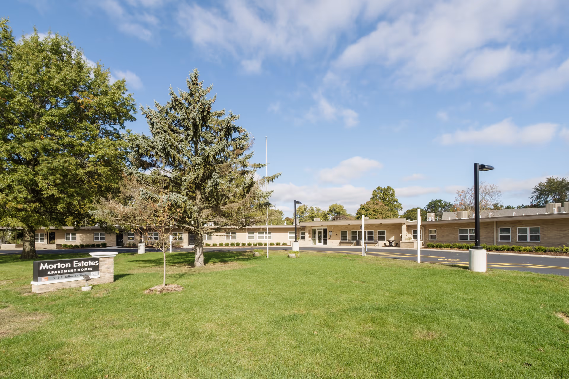 Single-story brick Morton Estates senior living building with a green lawn, trees, entrance canopy, and a sign in the foreground.