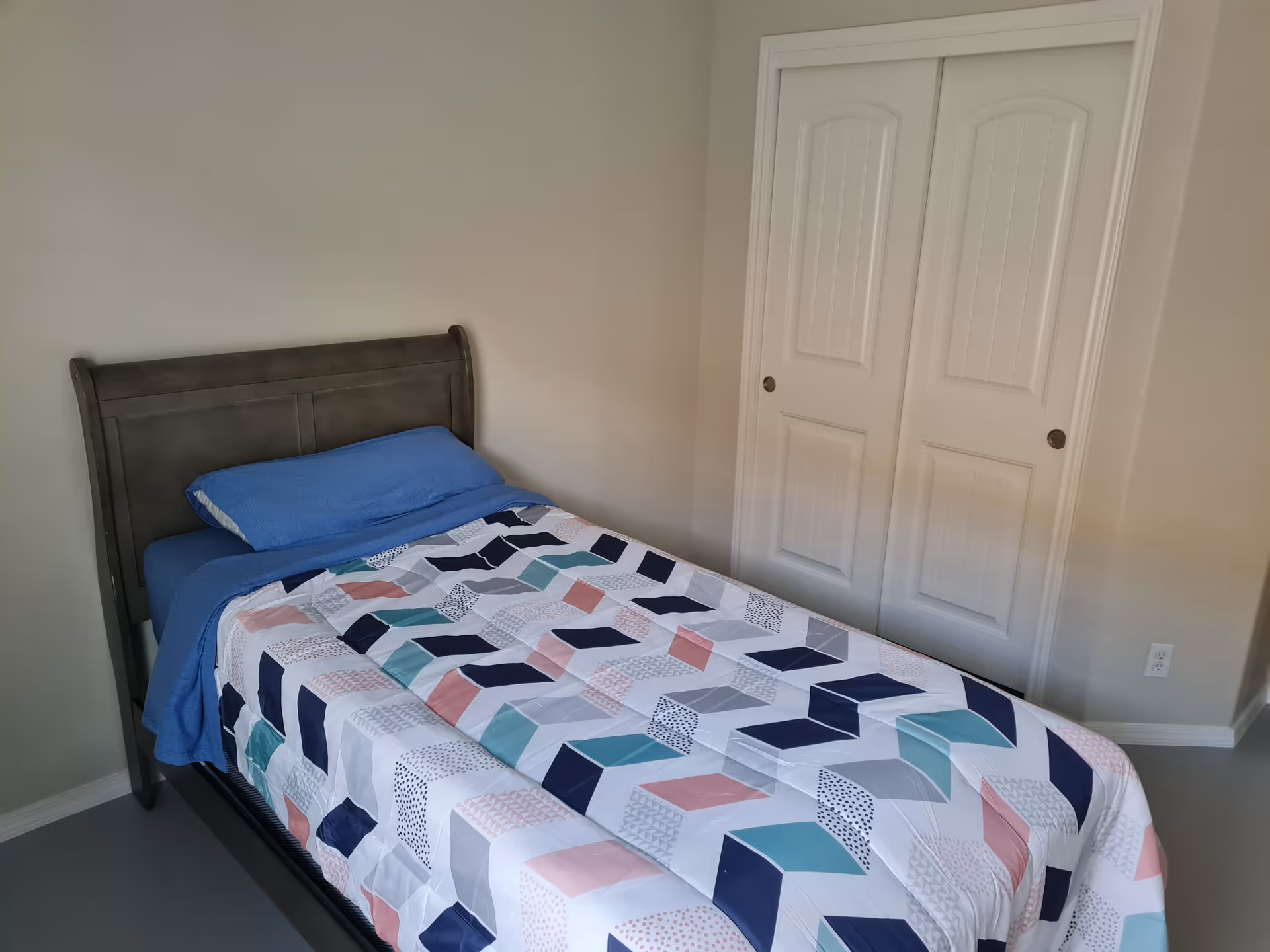 A simple bedroom with a twin bed covered by a geometric-patterned quilt and a blue pillow, next to white double closet doors.