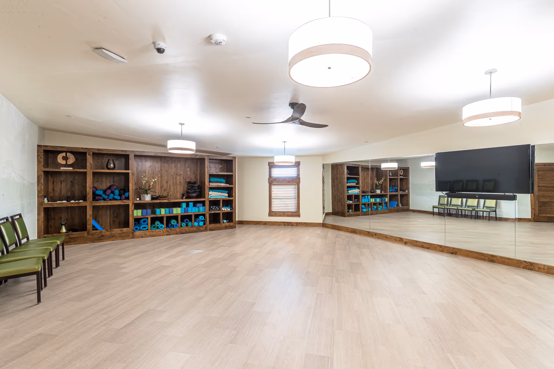 A spacious, well-lit exercise or yoga room with wooden flooring, large wall mirrors, and built-in wooden shelves stocked with yoga mats, blocks, and other exercise equipment. There are green chairs lined up against one wall and modern ceiling lights and a ceiling fan overhead.