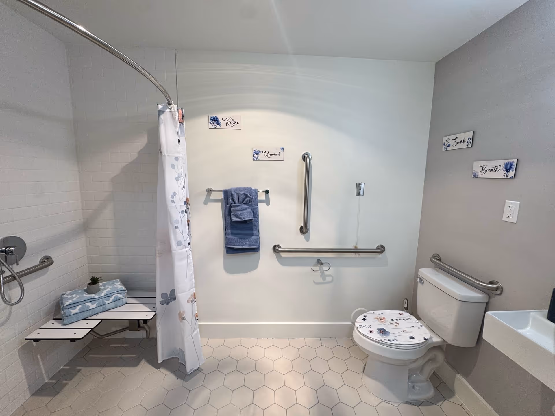 Accessible bathroom with a white tiled shower area featuring a foldable bench and grab bars. A shower curtain with a floral pattern is partially drawn. The bathroom has a white toilet with a floral-patterned seat cover, grab bars on the walls, a small white sink, and blue towels hanging on a towel rack. The walls have decorative signs with words like 'Relax', 'Unwind', 'Soak', and 'Breathe'. The floor is covered with hexagonal tiles.