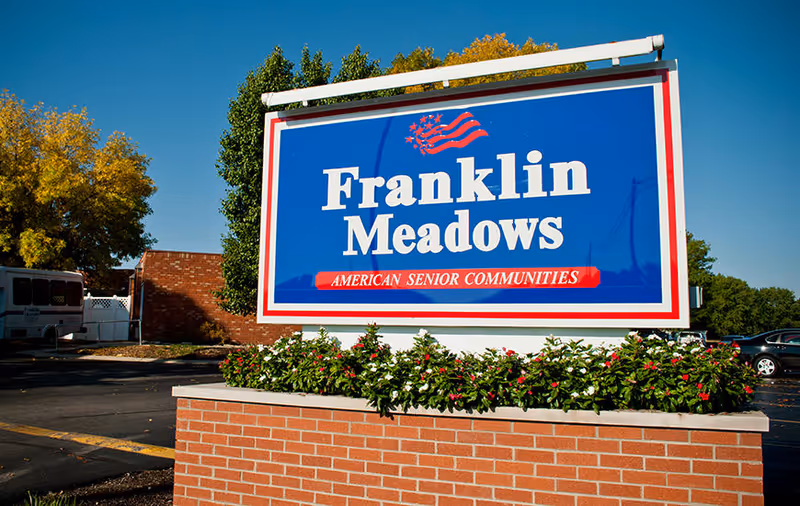 Outdoor sign for Franklin Meadows American Senior Communities with a blue background, white and red text, and an American flag graphic, set against a backdrop of trees and a clear blue sky.
