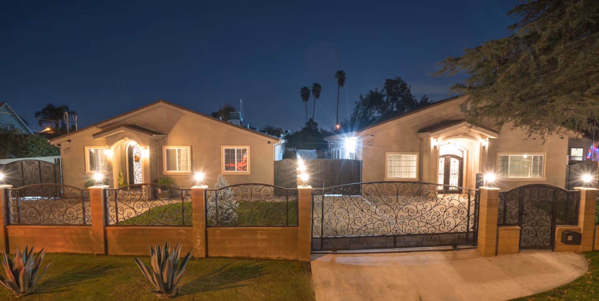 Nighttime view of two single-story homes behind a decorative wrought-iron front gate and low brick wall with exterior lights illuminating the entrances.