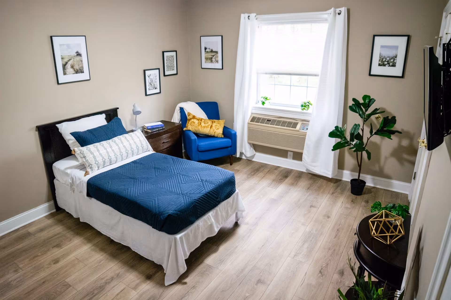 A neatly arranged bedroom in an assisted living facility featuring a single bed with blue bedding and white pillows, a wooden nightstand with a lamp and books, a blue armchair with a yellow cushion, a window with white curtains and an air conditioning unit below it, several framed pictures on beige walls, and wooden flooring with some green plants and decorative items.
