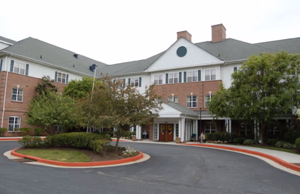 Front exterior view of a senior living facility named Charter Senior Living of Towson, featuring a three-story brick and white siding building with a gray roof, surrounded by trees and landscaping, and a circular driveway in front.
