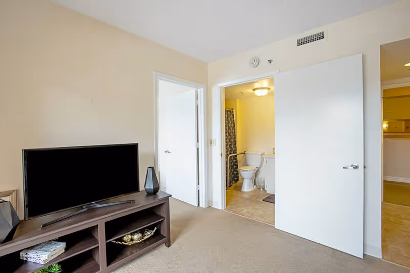 Living room with a TV on a media console and an open door revealing a bathroom with a toilet.