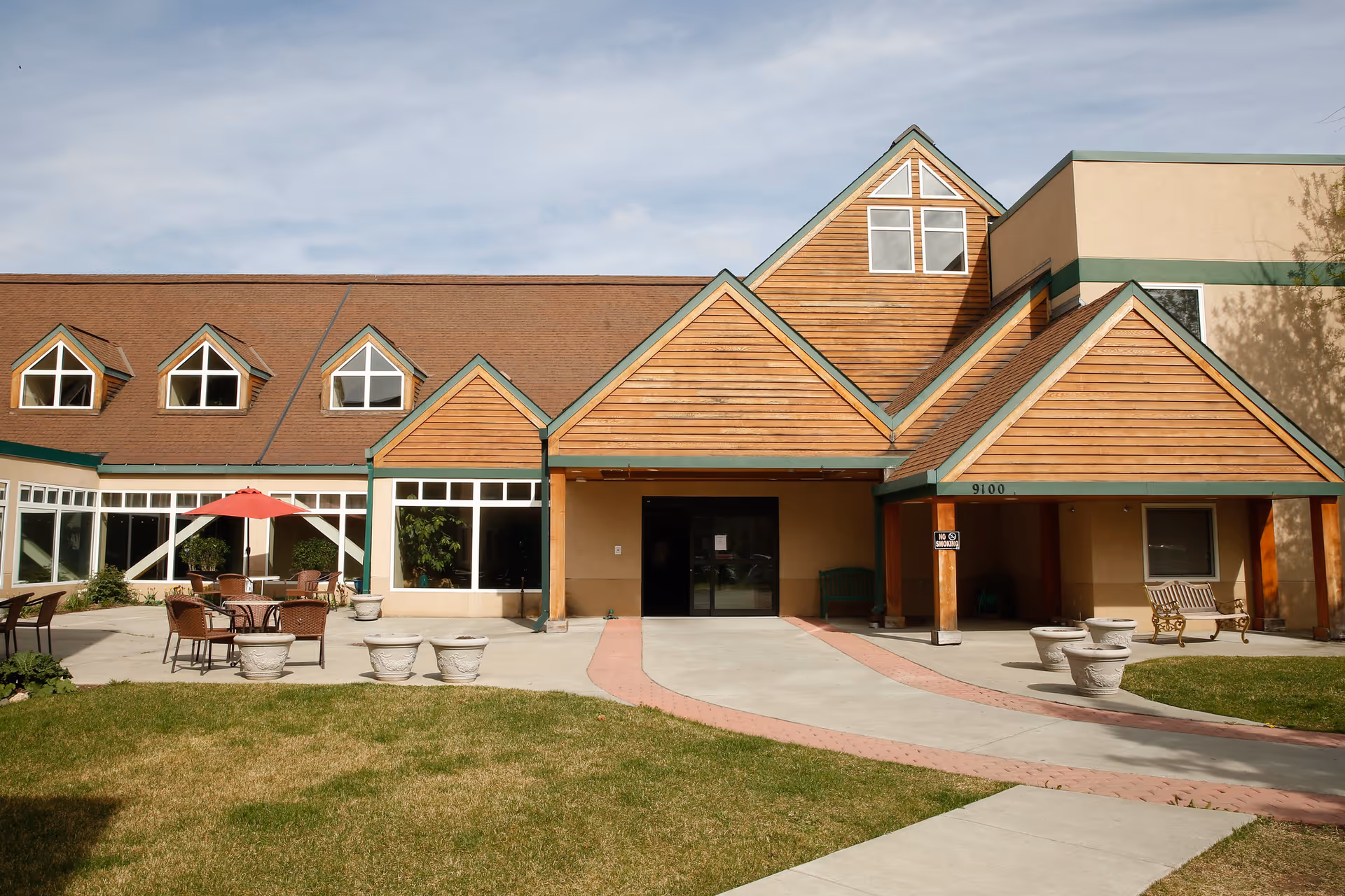 Front entrance of a senior living facility with a wooden gabled facade, courtyard seating and a red umbrella.