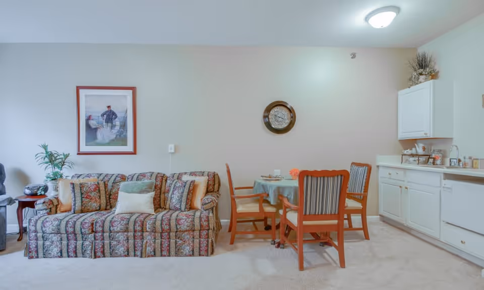 A cozy living area featuring a patterned sofa with multiple cushions, a small round dining table with four wooden chairs, and a kitchenette with white cabinets and a countertop. The wall behind the sofa has a framed painting, and there is a decorative plate hanging on the wall above the dining table. The room is carpeted and softly lit.