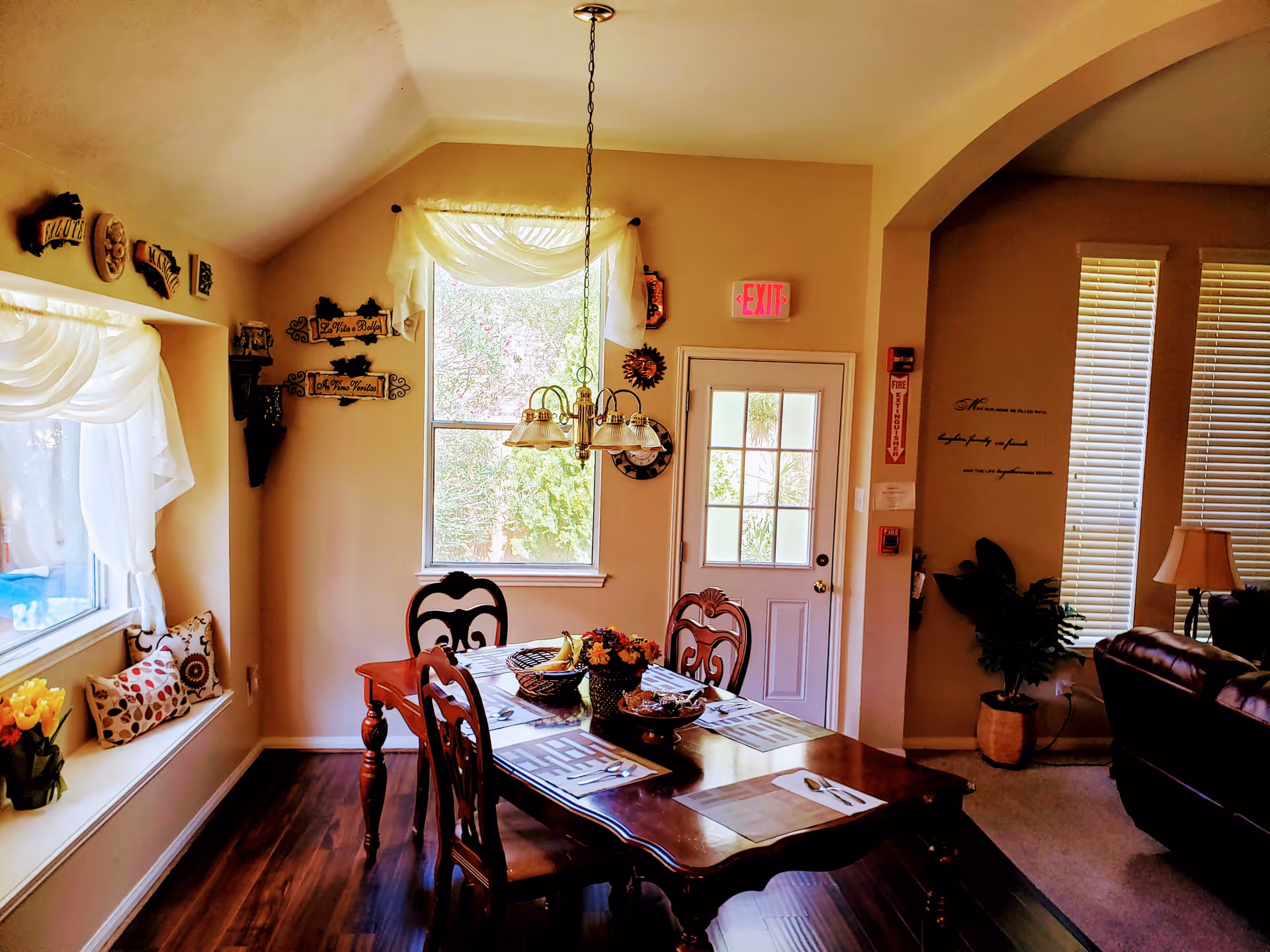 A cozy dining room with a wooden dining table set for four, featuring placemats, utensils, and a centerpiece with flowers and fruit. The room has a large window with sheer white curtains, a door with glass panes, and warm beige walls decorated with various wall hangings and an exit sign above the door. There is a hanging light fixture above the table and a glimpse of a living area with a leather chair and a lamp to the right.