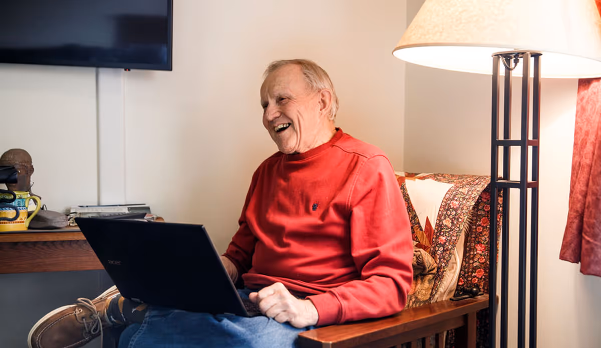 An elderly man wearing a red sweater and blue jeans is sitting on a wooden chair with a quilted cushion, using a laptop and smiling. There is a floor lamp with a white shade next to him, a television mounted on the wall, and a wooden table with various items in the background.