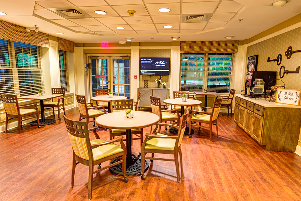 Well-lit communal dining room with round tables and wooden chairs, a TV on the far wall, and a buffet counter with a water dispenser.