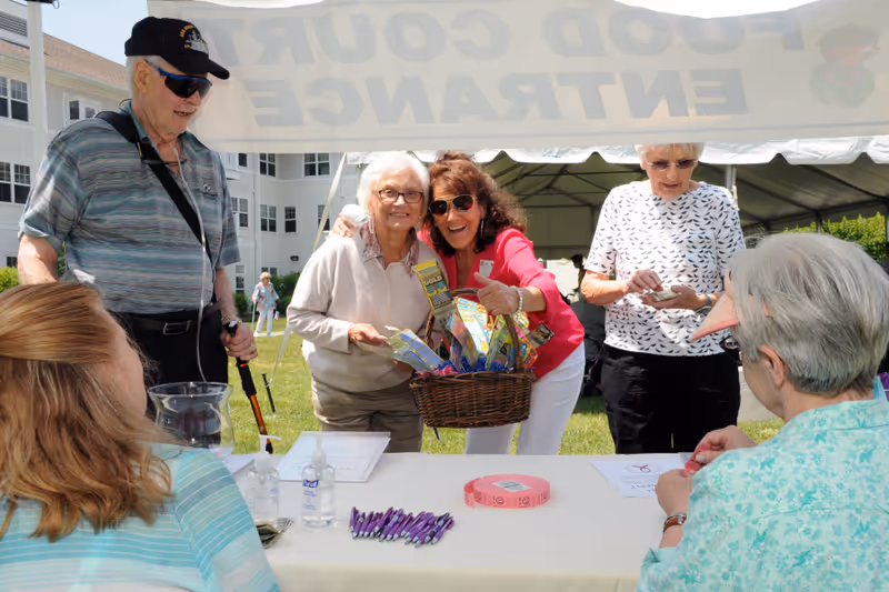 A group of elderly people and a staff member gathered outdoors at a food court entrance event. Two women stand behind a table with pens, hand sanitizer, and raffle tickets, while another woman in a red shirt holds a basket of snacks and poses with an elderly woman. A man with a cane and sunglasses stands nearby, and another elderly woman counts money. The setting is a grassy area with a large white tent and a multi-story building in the background.