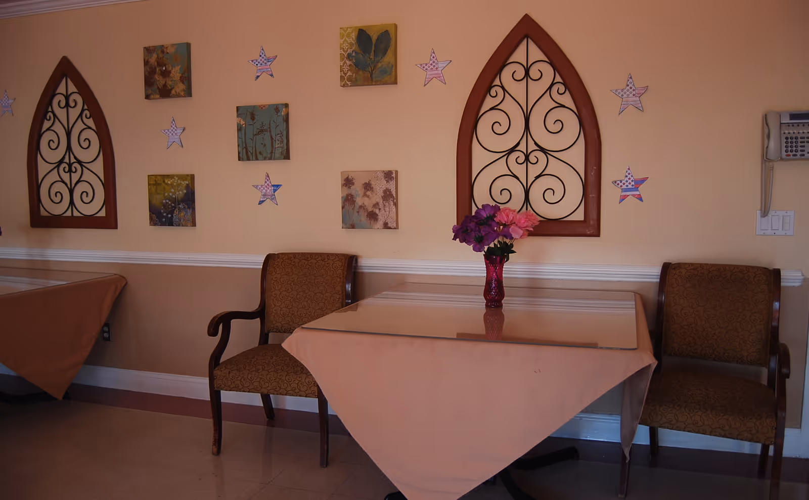 A dining area with two tables covered in peach-colored tablecloths, each accompanied by two upholstered chairs. On one table, there is a vase with purple and pink flowers. The wall behind the tables is decorated with framed artwork and star-shaped decorations, along with two decorative wrought iron wall hangings in wooden frames. A wall-mounted telephone and light switches are visible on the right side.