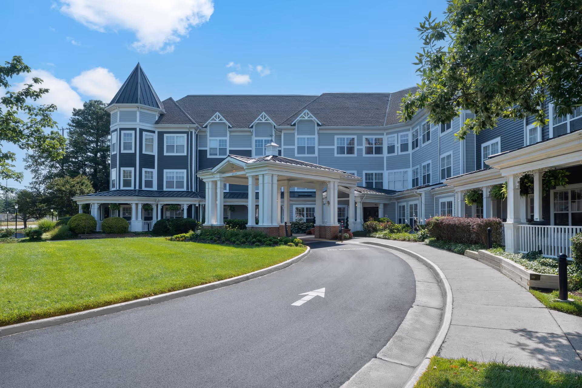 Exterior view of a senior living facility named Sunrise of Springfield featuring a large, multi-story building with gray siding and white trim. The building has a covered entrance with columns and a circular driveway with an arrow painted on the asphalt. There is a well-maintained lawn and landscaping with bushes and trees under a partly cloudy blue sky.