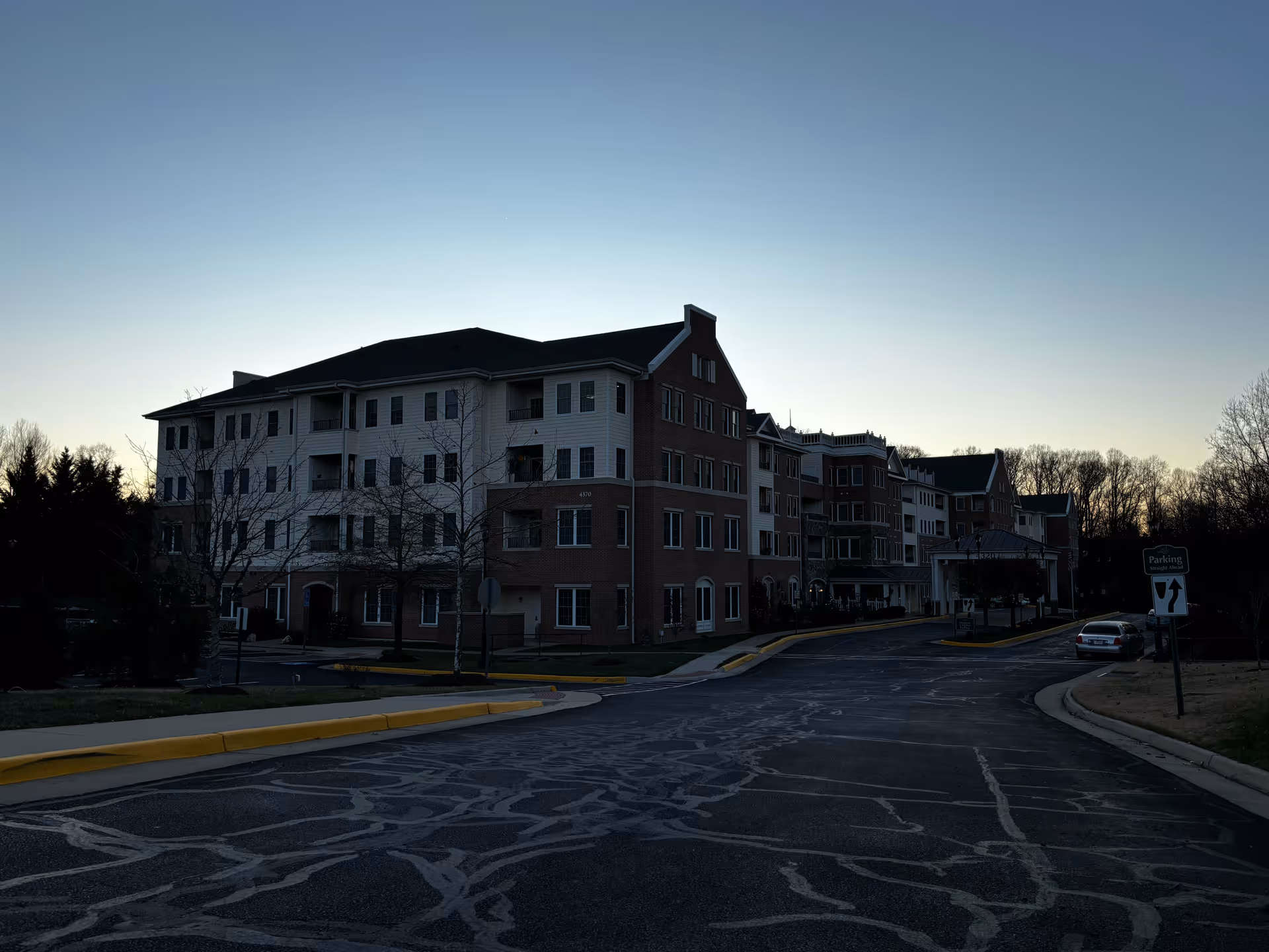 Front exterior of a multi-story senior living building at dusk with a curved driveway and a parking sign.