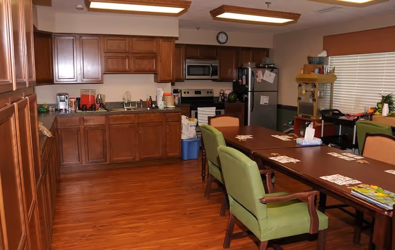 A kitchen and dining area with wooden cabinets, a stainless steel refrigerator, microwave, and stove. There is a long wooden table with green and orange cushioned chairs around it. The table has playing cards and tissues on it. The room has wooden flooring and a window with blinds on the right side.