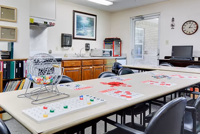 A room with a long table set up for a bingo game, featuring bingo cards, chips, and a bingo cage. The room has wooden cabinets, a popcorn machine, a microwave, a computer on a desk, a wall clock, and a framed picture on the wall. There is a door with a window leading outside.