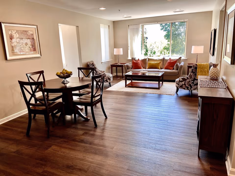 Bright open living area with a sofa, armchairs, coffee table and a round dining table on wood floors by a large window.