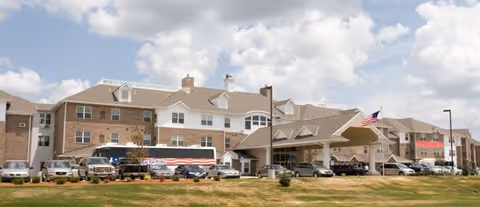 Exterior view of a large multi-story retirement resort building with a covered entrance, several parked cars, and a bus in front. The building has a mix of brick and white siding with multiple windows and a flagpole displaying the American flag.