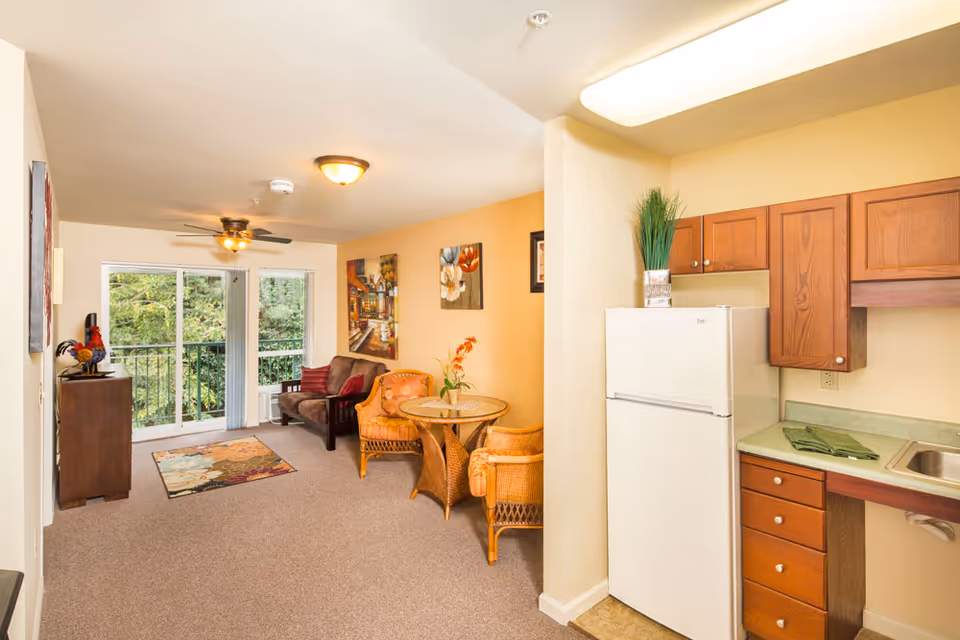 A cozy living area in a senior living facility featuring a small kitchen with wooden cabinets and a white refrigerator on the right. The living room has a brown loveseat, a round glass-top table with two wicker chairs, colorful wall art, and a ceiling fan. Large sliding glass doors lead to a balcony with a view of green trees outside.