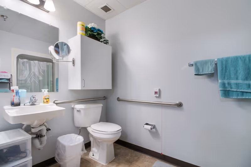 A clean and accessible bathroom with a white toilet, white sink, and a wall-mounted cabinet above the toilet. There are grab bars on the walls near the toilet and sink. A mirror is above the sink with a small round magnifying mirror attached to the wall. A towel rack with teal towels is mounted on the right wall. The floor has a light brown tile, and there is a small trash can next to the sink.