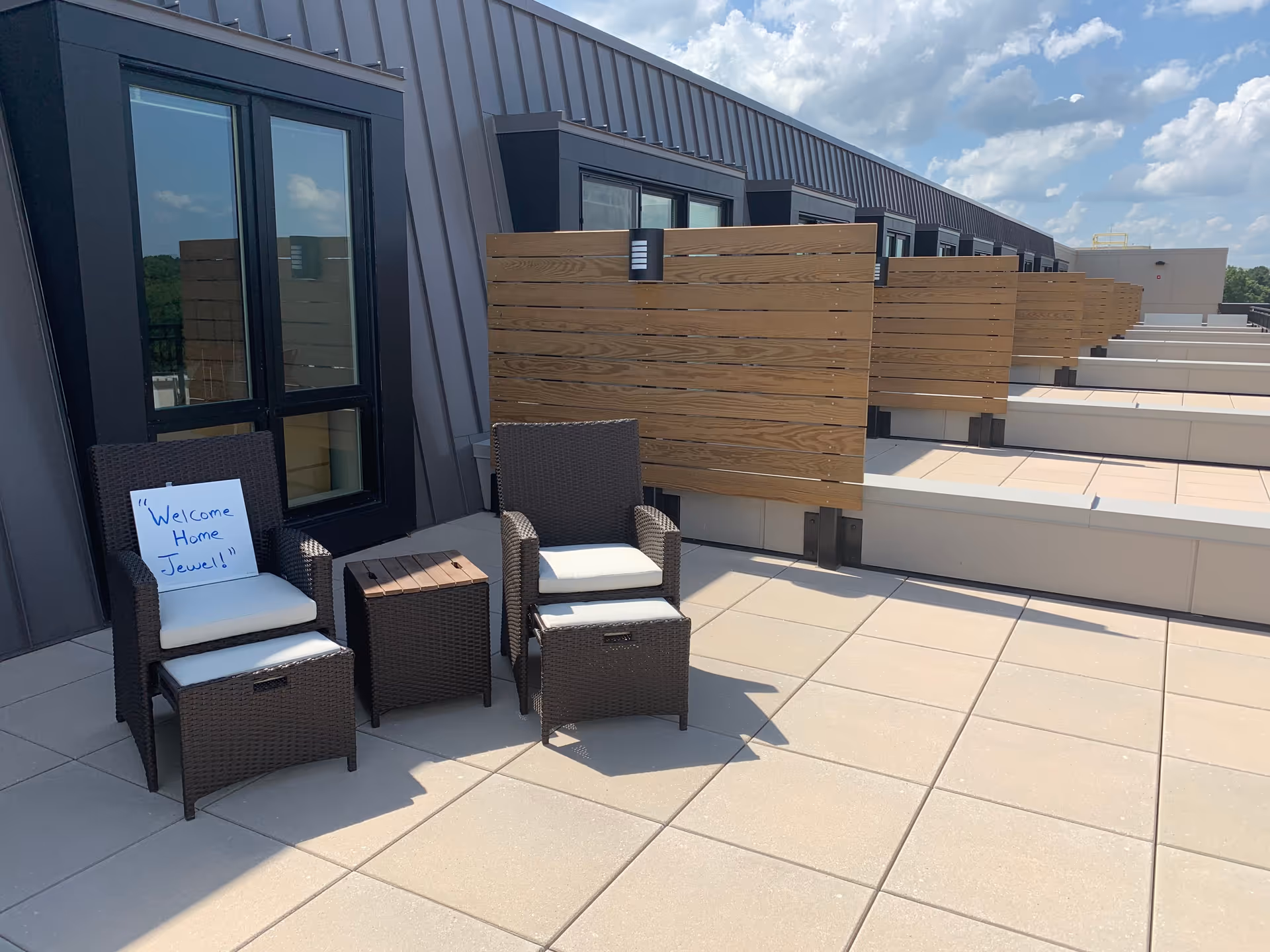 Rooftop patio with two wicker chairs, a small side table, wooden privacy screens, and a handwritten sign reading "Welcome Home Jewel!"