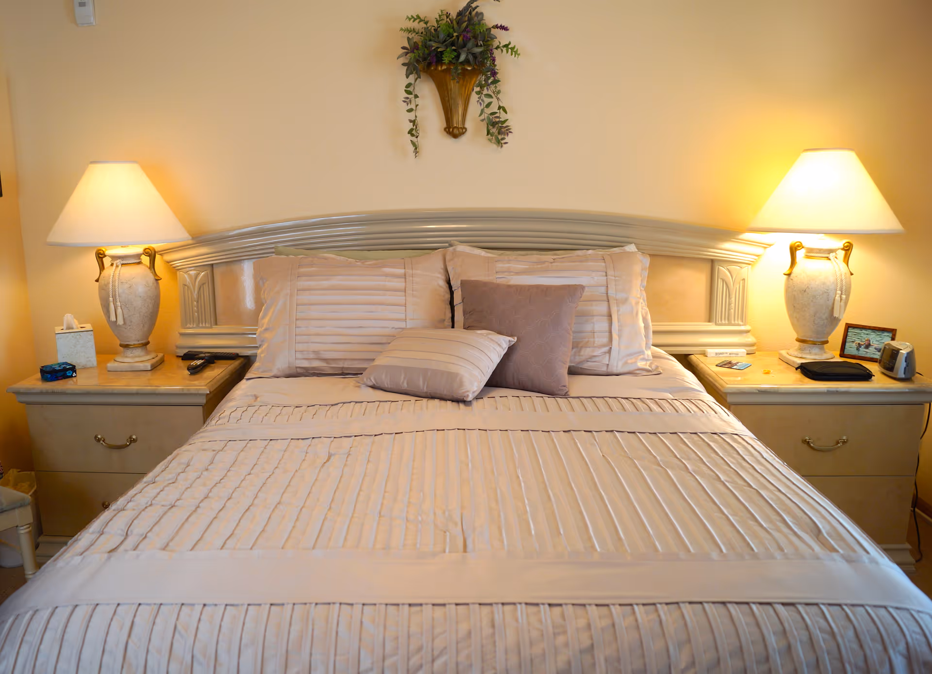 A neatly made bed with beige and light brown striped bedding and two pillows in front. On either side of the bed are matching nightstands with decorative lamps, a tissue box, a remote control, and other small items. Above the bed is a wall-mounted decorative plant holder with greenery.