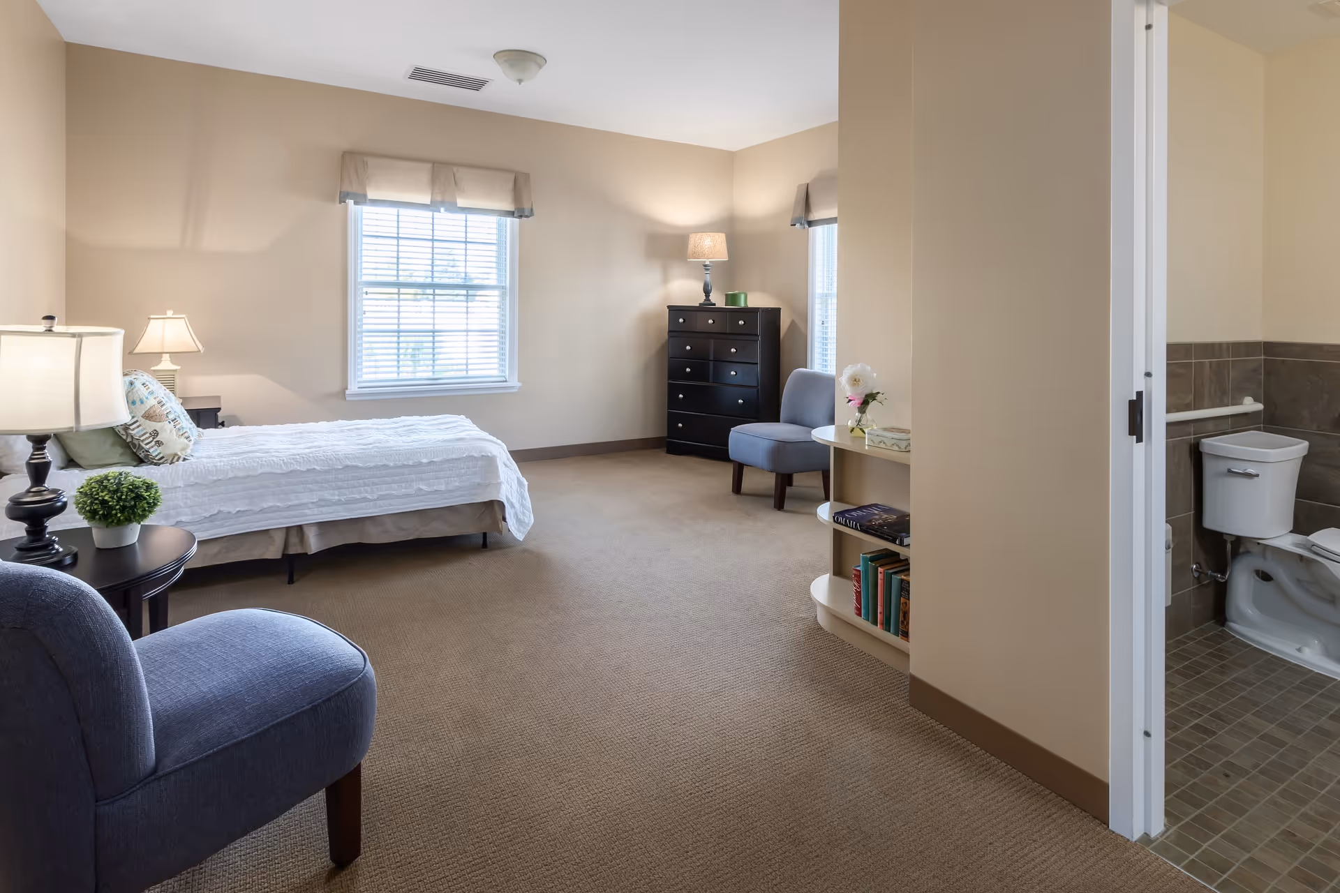 A bright and spacious bedroom in CountryHouse Residence featuring a single bed with white bedding, two blue upholstered chairs, a black dresser with a lamp, and a small round table with books and a flower vase. There are two windows with beige valances letting in natural light. An adjacent bathroom with a toilet and grab bars is visible through an open door.